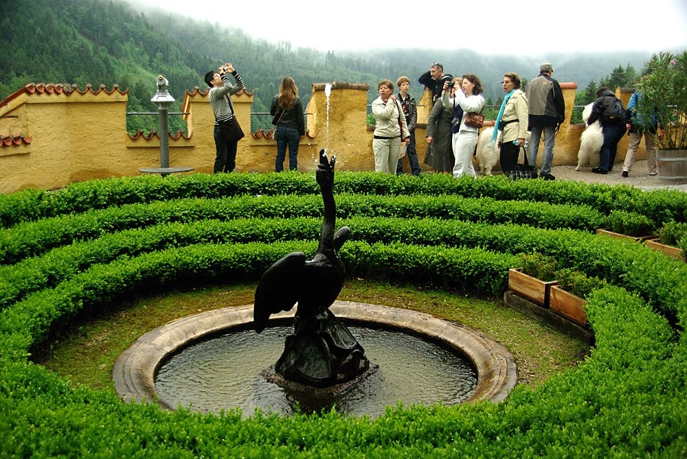 Beautiful swan fountain at Hohenschwangau castle surrounded by green bushes and visiting tourists.