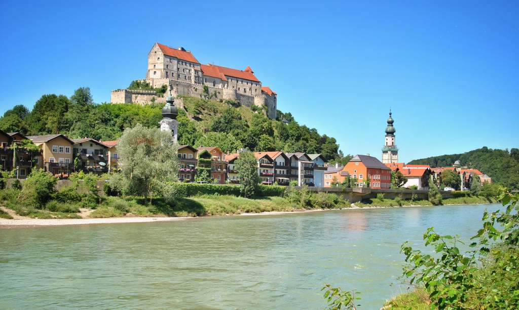 A beautiful view of Burghausen Castle from the Salzach river surrounded by houses and trees and other greenery.