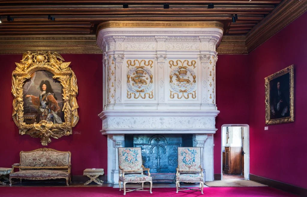 An elegant looking interior inside the Château de Chenonceau with red wallpaper and hanging portraits.